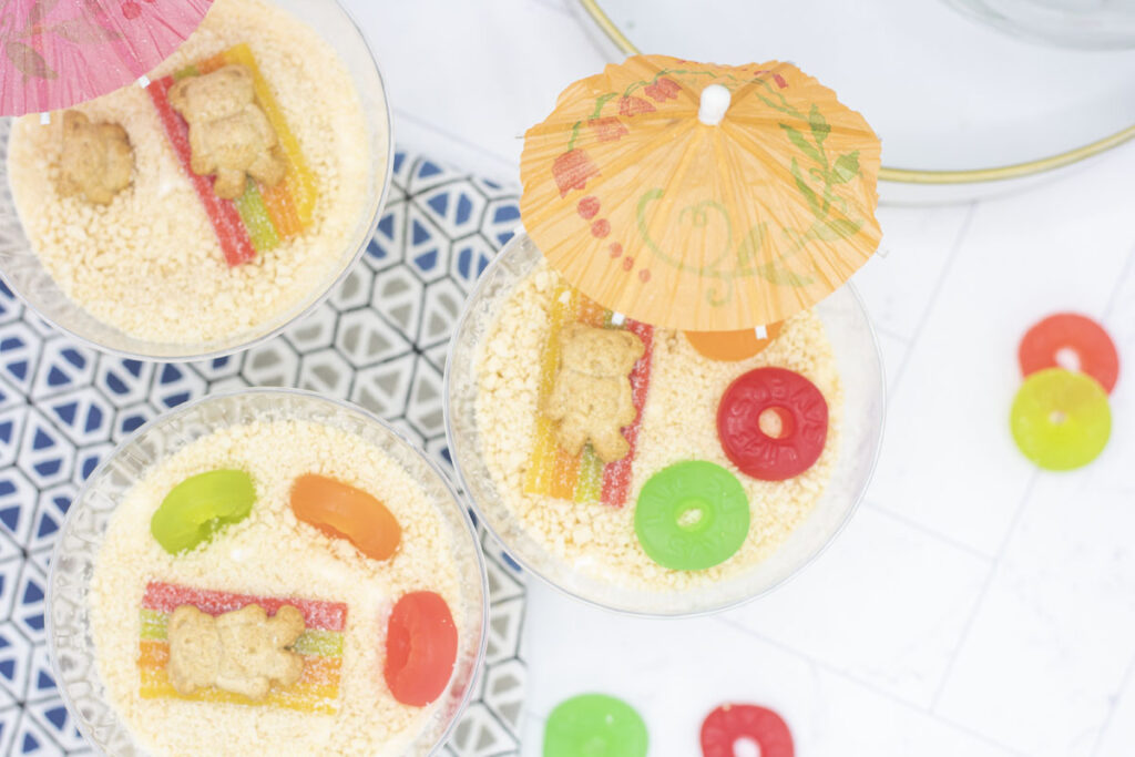 Vanilla dirt cups with a beach scene: teddy grahams on a rainbow sour strip beach towel surrounded by lifesavers