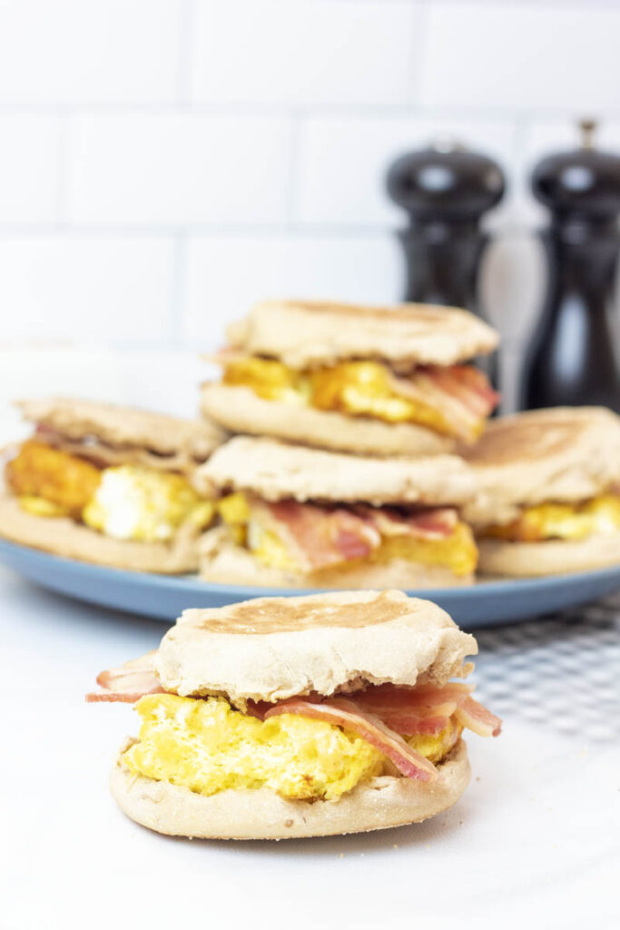 A plate of breakfast sandwiches behind a single bacon egg and cheese on an english muffin