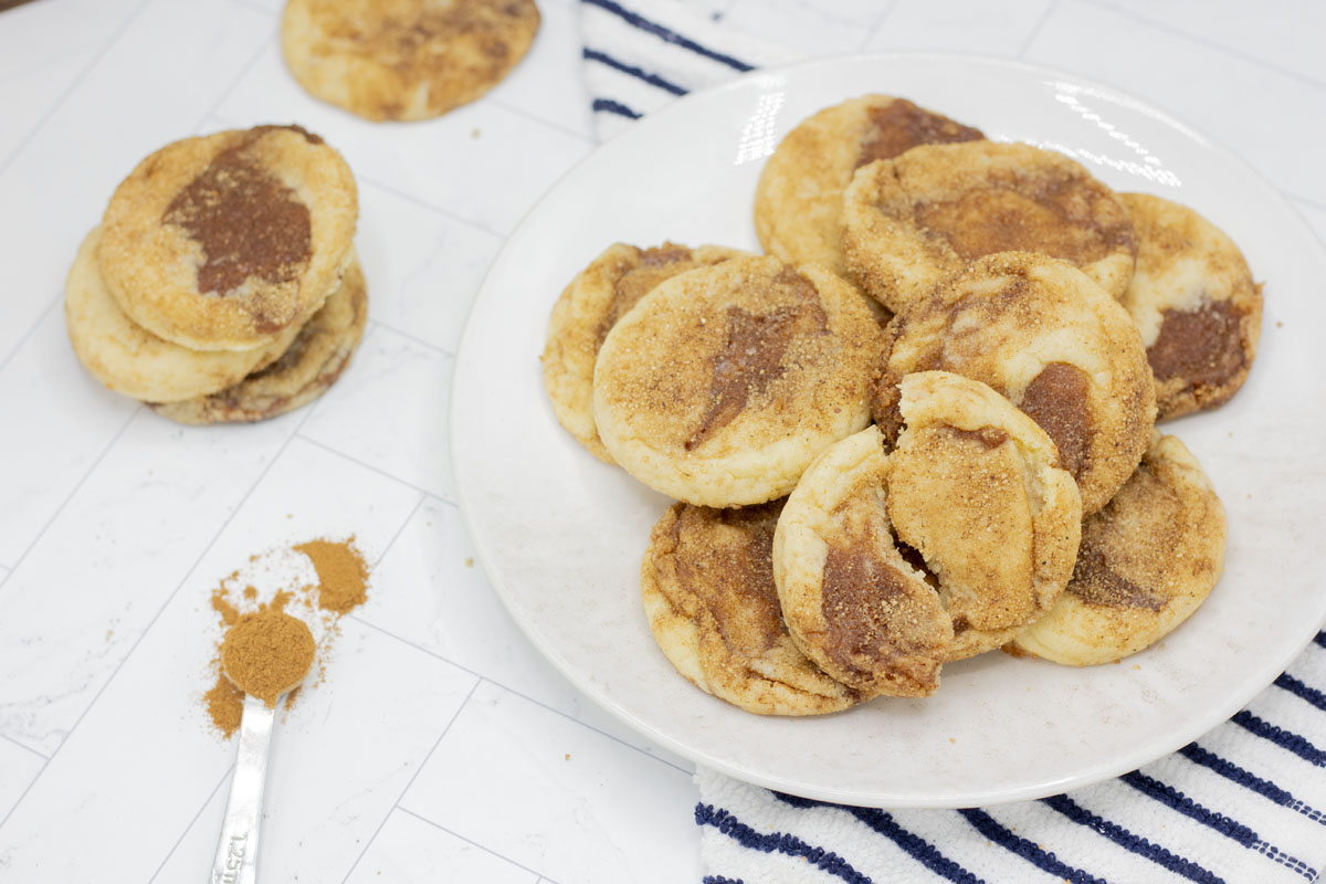 A plate of cinnamon roll snickerdoodles, with cinnamon and additional cookies nearby