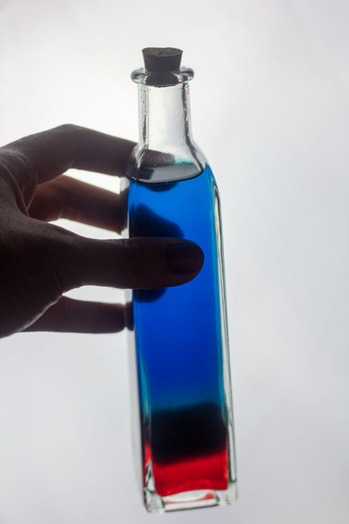 Hand holding a blue cocktail that fades to red in a tall square potion bottle