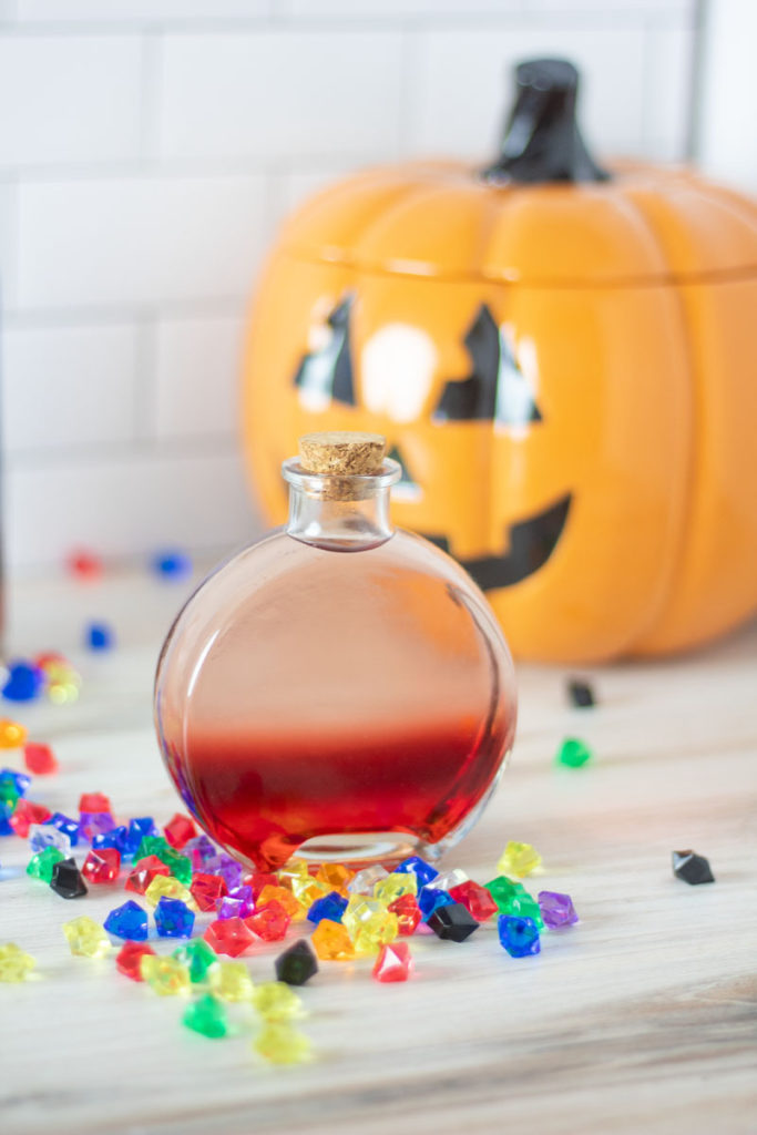 A red and clear ombre cocktail in a potion bottle with gems scattered around and a pumpkin