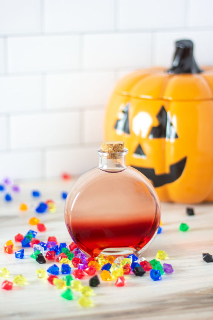A red and clear ombre cocktail in a potion bottle with gems scattered around