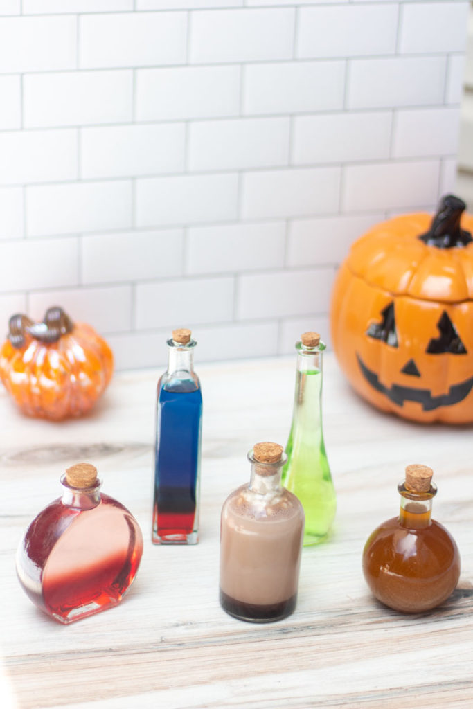 Five potion bottles containing various Halloween cocktails with pumpkins in the background