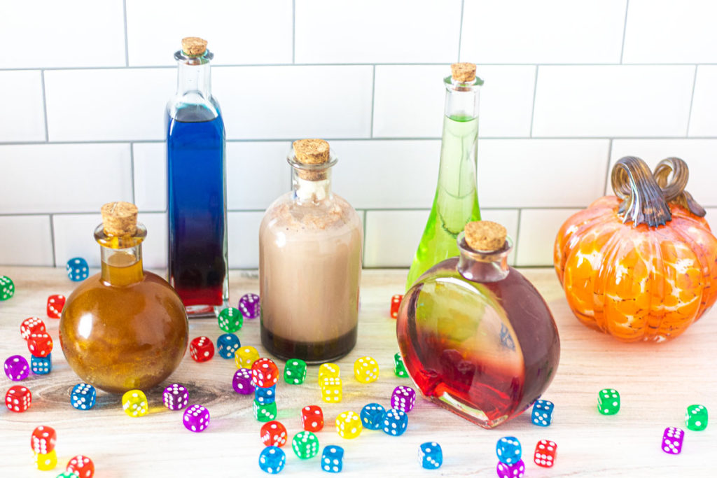 Five halloween cocktails in different potion bottles with dice and pumpkins scattered around