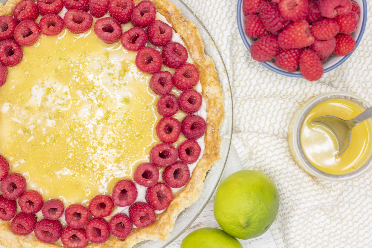 Top down view of a raspberry lime cheesecake surrounded by limes, citrus curd, and raspberries in a bowl