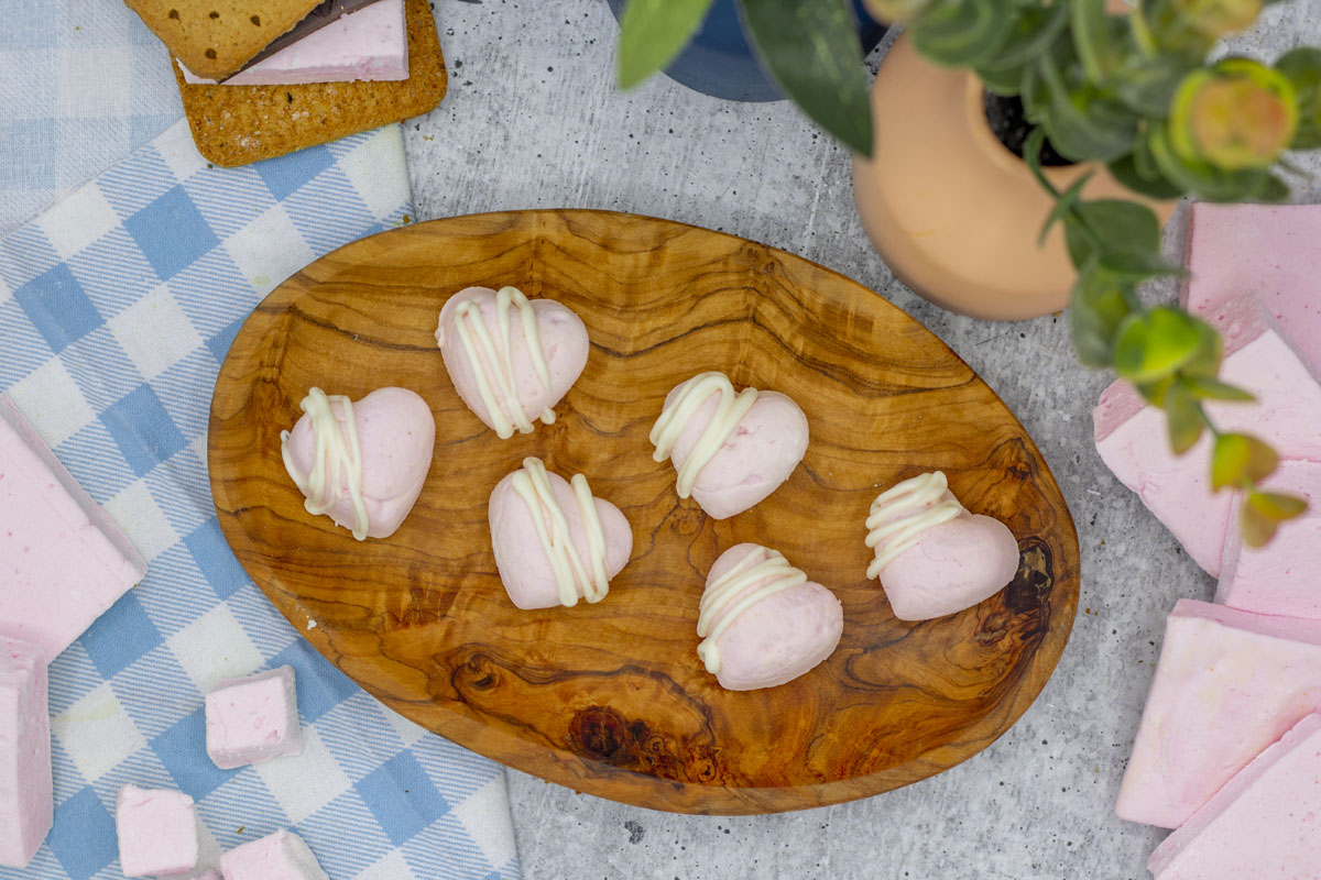 Heart shaped raspberry marshmallows drizzled with white chocolate on a small wooden plate