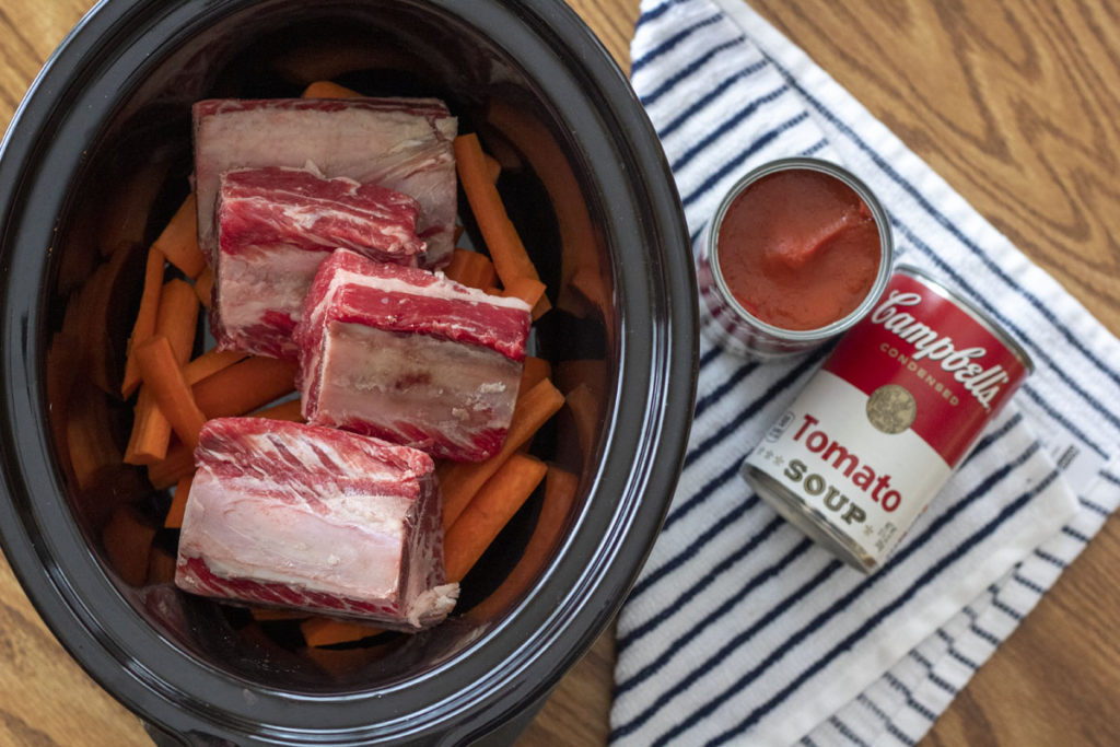 Crock pot filled with short ribs and carrots beside tomato soup cans for slow cooker short ribs