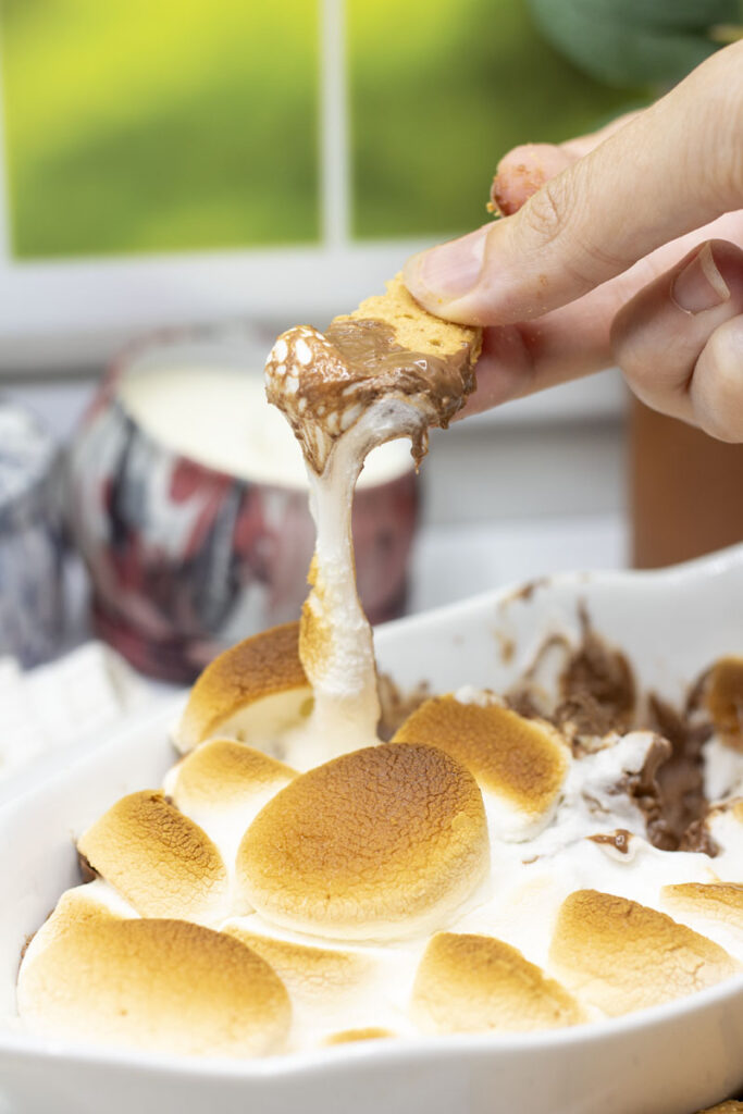 Graham cracker dipped in s'mores dip with stringy melted marshmallow pulling from the graham cracker