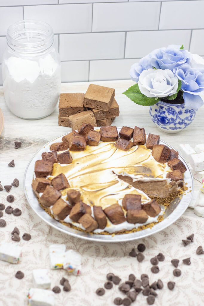 S'mores french silk pie with a slice missing surrounded by scattered chocolate chips, chocolate marshmallows and marshmallow fluff