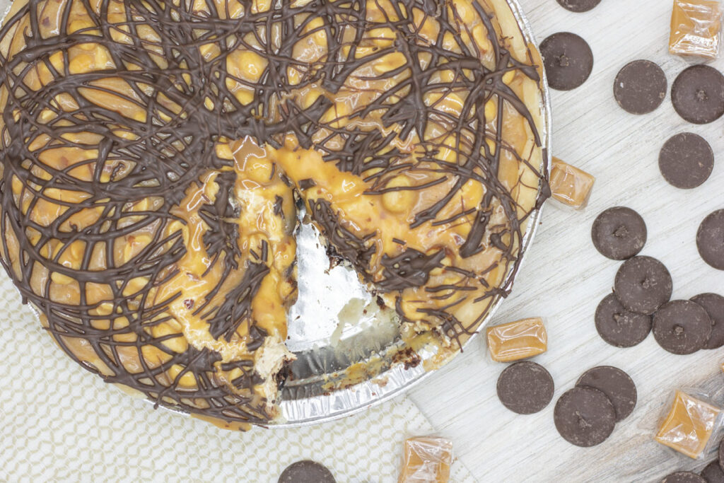 View of snickers peanut butter pie with a slice cut out and chocolate and caramels scattered around