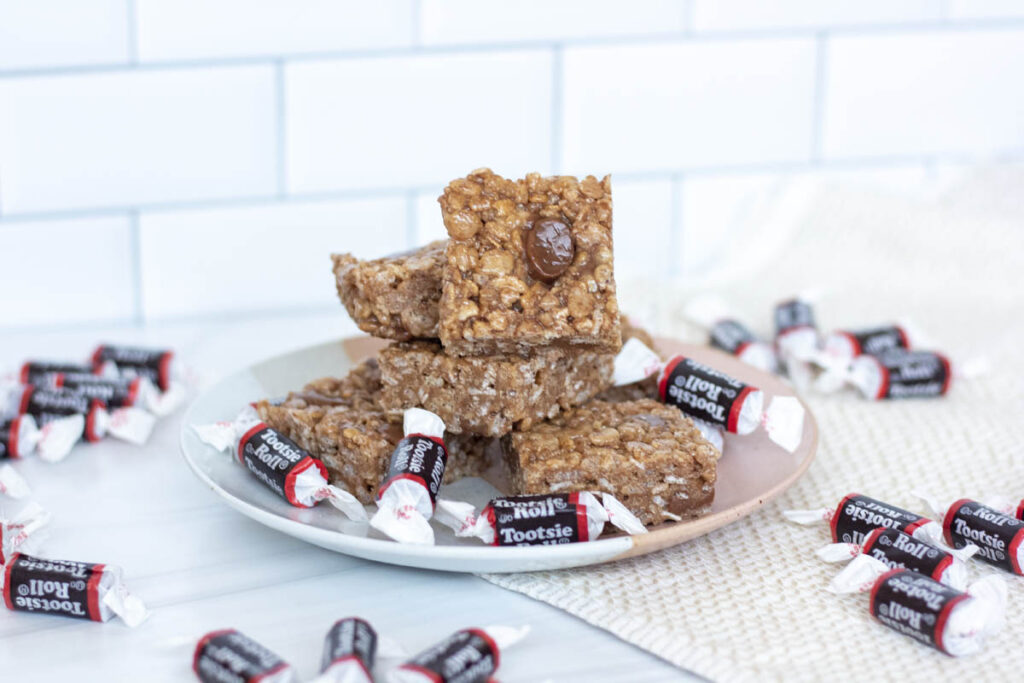 Assorted tootsie roll rice krispy treats stacked on a plate with scattered tootsie rolls
